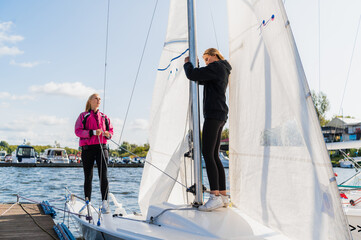 Two young female athletes prepare a sailing boat for the regatta on the river pier.