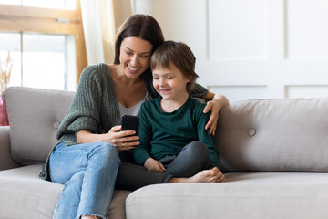 Loving young Caucasian mom and small son relax on couch in living room watch video on smartphone together. Happy caring mother or nanny and little boy child rest on sofa using modern cellphone.