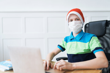 Serious boy in a disposable face mask and Santa Claus hat during online classes