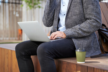 Cropped shot of busy elegant mature man talking on the phone, working on his laptop while sitting on the bench outdoors