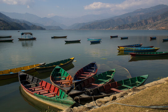 Colorful boats floating with no people anchored on the Phewa Lake, Pokhara, Nepal. Nice reflection of the sky and clouds on the water. Large mountains far away on the background. Post pandemic travel