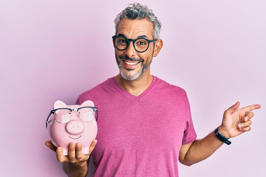 Middle age grey-haired man holding piggy bank with glasses smiling happy pointing with hand and finger to the side