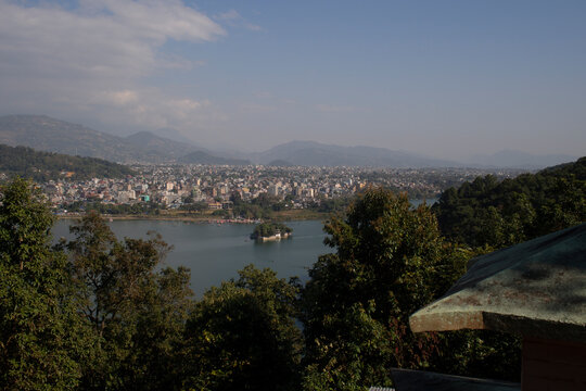 Beautiful panoramic view of Phewa lake and the city at a distance. Trees and a roof in the foreground and mountain chain in the background, some clouds in the sky of Pokhara, Nepal.