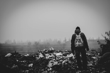 A man in old clothes and with a backpack stands against the background of a landfill.
