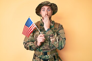 Young caucasian man wearing camouflage army uniform holding usa flag covering mouth with hand,...