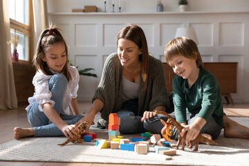 Fototapeta premium Caring single mother have fun play toys with excited two small kids at home. Happy loving young Caucasian mom engaged in game activity together with little children, enjoy family weekend indoors.
