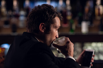 a young man drinks a cocktail from a glass in a bar, holding a smartphone, close-up.