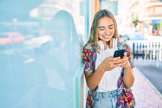 Young beautiful blonde caucasian woman smiling happy outdoors on a sunny day wearing headphones and using smartphone