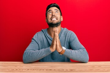 Fototapeta premium Handsome hispanic man wearing casual style sitting on the table begging and praying with hands together with hope expression on face very emotional and worried. begging.