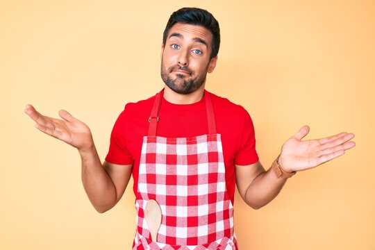 Young hispanic man wearing professional baker apron clueless and confused with open arms, no idea and doubtful face.