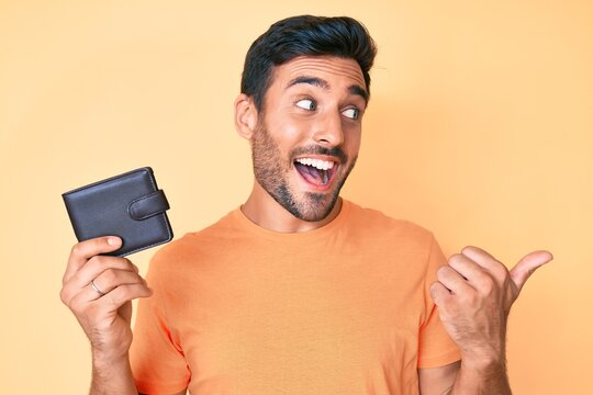 Young hispanic man holding leather wallet pointing thumb up to the side smiling happy with open mouth