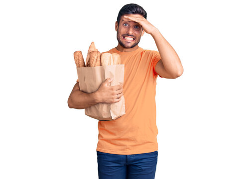 Young hispanic man holding paper bag with bread stressed and frustrated with hand on head, surprised and angry face