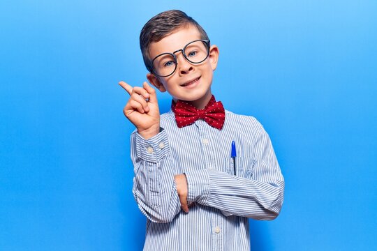 Cute blond kid wearing nerd bow tie and glasses with a big smile on face, pointing with hand and finger to the side looking at the camera.
