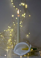 white burning candle, beautiful and noble white callas flower, illuminated background of a chain of white lights, Christmas waiting time, winter