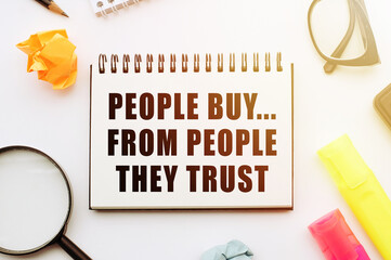 Text - People Buy From People They Trust in notebook on white table with office tools.