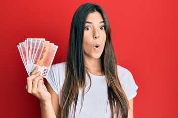 Young brunette woman holding 100 norwegian krone banknotes scared and amazed with open mouth for...
