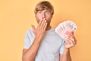 Young blond man holding israeli shekels banknotes covering mouth with hand, shocked and afraid for...