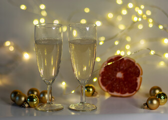 two glasses of champagne, pink grapefruit, golden glass balls and various Christmas decorations, illuminated background of a chain of white lights, Christmas waiting time