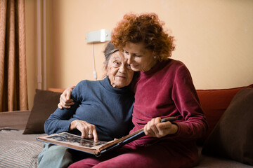 Positive aged ladies looking album photos sitting sofa at home, cheerful friends. Senior woman and her mature nurse watching photo album. Granny showing her daughter memories from the past
