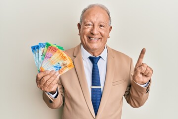 Senior caucasian man holding swiss franc banknotes smiling with an idea or question pointing finger with happy face, number one