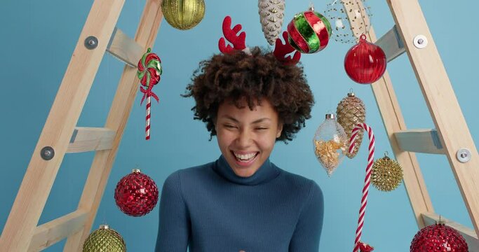 Slow motion of overjoyed dark skinned woman laughs happily hears funny anecdote from friend has fun on New Year party poses against shiny toys for fir tree cannot stop laughing. Holiday celebration