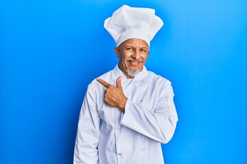 Middle age grey-haired man wearing professional cook uniform and hat cheerful with a smile of face pointing with hand and finger up to the side with happy and natural expression on face