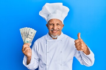 Middle age grey-haired man wearing professional cook uniform holding usa dollars banknotes smiling happy and positive, thumb up doing excellent and approval sign
