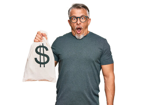 Middle age grey-haired man holding dollars bag scared and amazed with open mouth for surprise, disbelief face