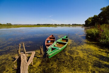 countryside landscape with little shabby worn wooden fishing boats at a lake bank footbridge in rich green vegetation, swamp covered with duckweed, ecology tourism concept