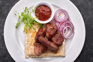 shashlik, with sauce, onions, and lavash, on a white plate, on a dark background