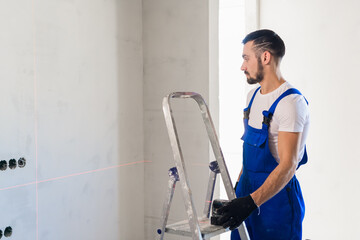 The handyman put the laser level on the ladder and looks at the markings on the wall