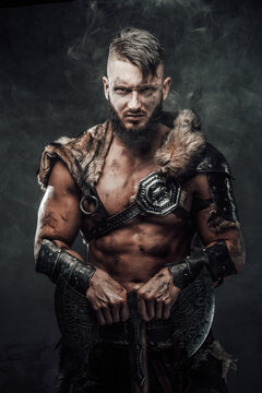 Savage and barbaric viking seafarer holding two handed axe in dark foggy background staring at camera.