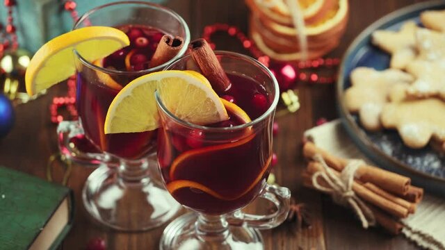Hand puts a cup of fragrant, hot wine with spices and fruits on the table with branches of a Christmas tree and glowing garland lights. Preparing and serving a hot Christmas drink.