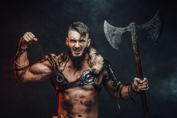 Nordic barbarian with lights armour showing his huge biceps looking at camera and holding his two handed axe in dark background.