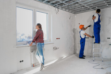 A man in a blue uniform with a drill stands on a stepladder. A bearded man in a hard hat holds a...
