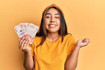 Young latin woman holding colombian pesos screaming proud, celebrating victory and success very...