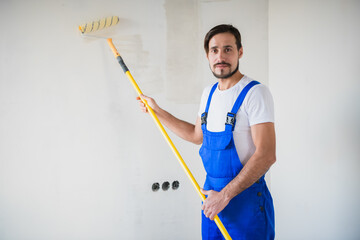 A builder in a blue overalls holds a roller in his hands, smiles and looks at the camera