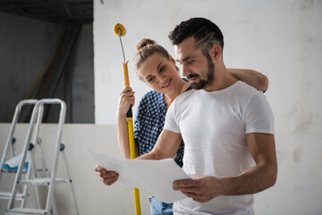 A man holds a schema of a house in his hands. A woman stands behind and holds a roller in her hands