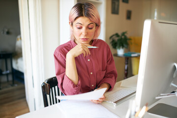 Serious young female employee with pink hair doing paperwork from home office, sitting at desk with...