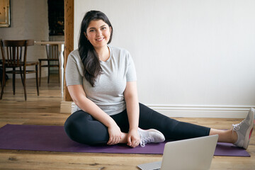 Indoor image of beautiful energetic young plus size woman with curvy body training at home to lose weight and get strong flexible body, wearing sneakers, exercising on mat, watching video on laptop