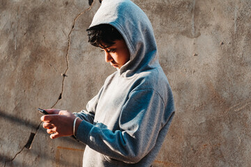 Portrait of little Indian school kid wearing hoodie using mobile phone at home. Concept of online...