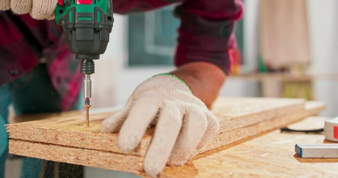 Concentrated young bearded carpenter wearing jeans and a plaid flannel shirt drilling a hole in a piece of wood with a portable electric drill in his carpentry workshop.