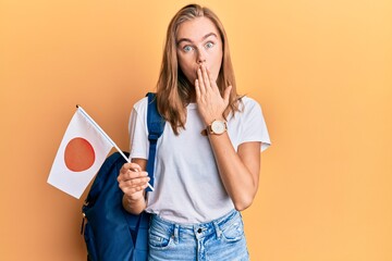 Beautiful blonde woman exchange student holding japan flag covering mouth with hand, shocked and...