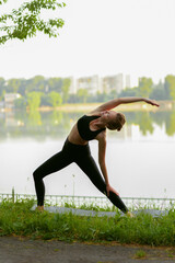 Fototapeta premium Full length image of a young woman in black sportwear doing yoga in the park, lake background.