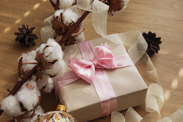 christmas gift in craft paper, with pink ribbon, pine cones and branches with cotton flowers on the wooden table
