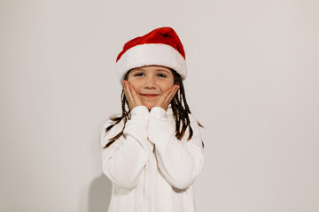 lovely happy little girl wearing white dress and santa hat touching her face and posing over isolated background