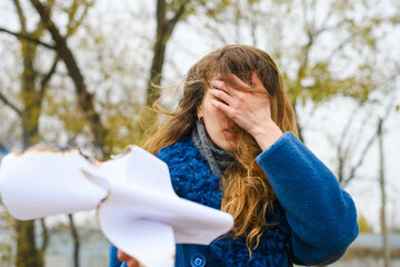 Young frustrated woman in blue coat cover face with hand and hold burning white blank paper sheet outdoors