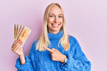 Young blonde girl holding 100 norwegian krone banknotes smiling happy pointing with hand and finger