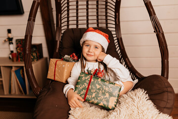 Indoor portrait of happy lovely little girl in dress and santa hat posing at camera with Christmas presents during holiday photoshoot