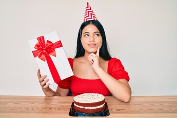Young asian girl wearing birthday hat holding present serious face thinking about question with...